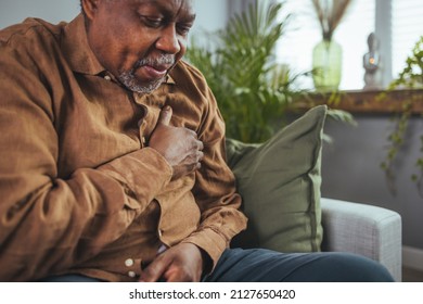 African Senior Man Sitting On Sofa At Home With Sickness. Elderly Male Suffering From Heart Attack And Feel Unwell Need For Medicine. Retirement People Health Care And Illness Recovery Concept