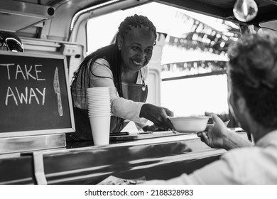 African Senior Woman Serving Takeaway Food Inside Food Truck - Focus On Chef Female Face - Black And White Editing