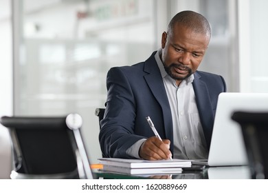 African Serious Businessman Writing Notes And Using Laptop. Mature Business Man Writing His Strategy In Modern Office. Focused Black Entrepreneur Sitting At Desk In Modern Office While Working.