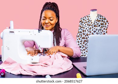 african tailor with afro dreadlocks pigtails sews clothes on sewing machine at tailor office pink wall background