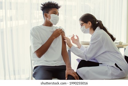 African Teen Man Wearing Face Mask, Getting Vaccination To Protect Or Prevent Covid19 Virus, Getting Scary Vaccine Injection While Female Caucasian Doctor Preparing Syringe To Vaccinate At Hospital