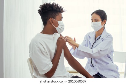 African Teen Man Wearing Face Mask, Getting Vaccination To Protect Or Prevent Virus, Getting Scary Of Vaccine Injection While Female Asian Doctor Preparing Syringe To Vaccinate Patient At Hospital