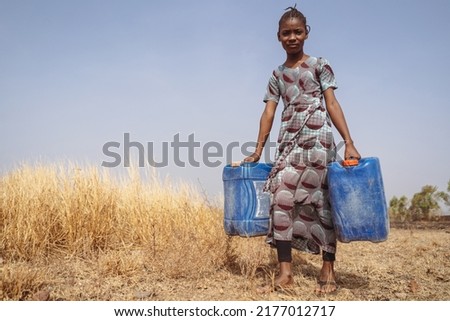 African teenager with two blue water cans walking through a field of dried up grass to get water from the public pump; global warming concept