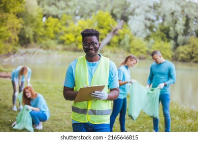 African Volunteer Man With Clipboard In Park. Africa Volunteering, Charity, People And Ecology Concept. Behind Him Are Volunteers Cleaning The Public Park.

