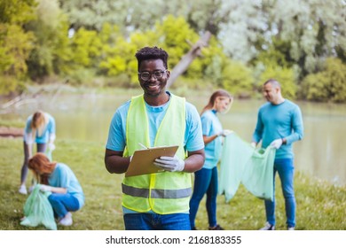 African Volunteer Man With Clipboard In Park. Africa Volunteering, Charity, People And Ecology Concept. Behind Him Are Volunteers Cleaning The Public Park.

