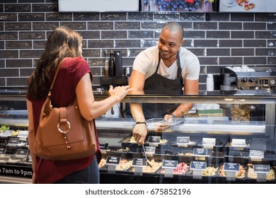 African waiter serving fresh food to young woman. Happy smiling guy preparing take away salad for woman customer. Black man with apron taking pasta salad in spoon and serving to customer. - Powered by Shutterstock