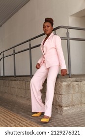 African Woman With Afro Hair Wearing Pink Summer Suit  Posing Outside At Urban Minimalist Background. Glamorous And Modern Style Runaway Full Length Portrait. Modeling And 2022 Fashion Concept.