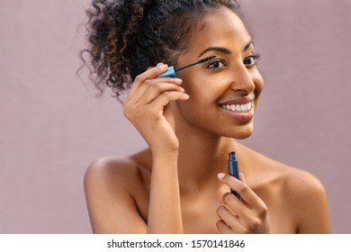 African Woman Applying Black Mascara On Eyelashes With Brush. Young Beautiful Woman Applying Makeup On Eyes. Portrait Of Black Beauty Girl Applying Makeup Isolated Over Pink Background With Copy Space