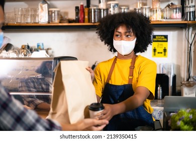 African Woman Barista  Wear Mask Due To Covid-19 Pandemic, Receive Drink On Coffee Bar Counter. Restaurant Worker Giving Takeaway Delivery Food Bag.Small Business Social Distance Concept.