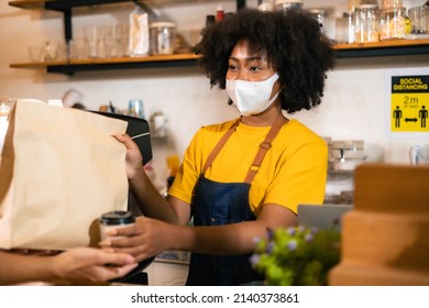 African Woman Barista  Wear Mask Due To Covid-19 Pandemic, Receive Drink On Coffee Bar Counter. Restaurant Worker Giving Takeaway Delivery Food Bag.Small Business Social Distance Concept.