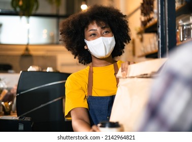 African Woman Barista  Wear Mask Due To Covid-19 Pandemic, Receive Drink On Coffee Bar Counter. Restaurant Worker Giving Takeaway Delivery Food Bag.Small Business Social Distance Concept.