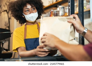 African Woman Barista  Wear Mask Due To Covid-19 Pandemic, Receive Drink On Coffee Bar Counter. Restaurant Worker Giving Takeaway Delivery Food Bag.Small Business Social Distance Concept.