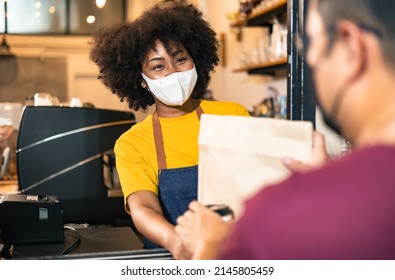 African Woman Barista  Wear Mask Due To Covid-19 Pandemic, Receive Drink On Coffee Bar Counter. Restaurant Worker Giving Takeaway Delivery Food Bag.Small Business Social Distance Concept.