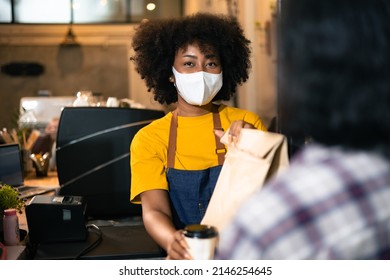 African Woman Barista  Wear Mask Due To Covid-19 Pandemic, Receive Drink On Coffee Bar Counter. Restaurant Worker Giving Takeaway Delivery Food Bag.Small Business Social Distance Concept.