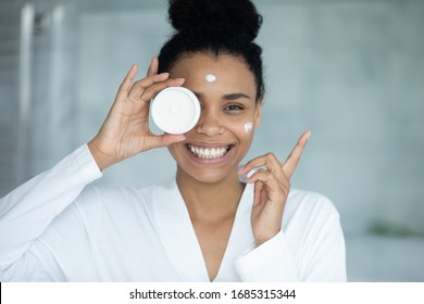 African woman in bathrobe applied cream on face holds jar feels happy looks at camera, anti-wrinkle treatment, remedy for complexion improvement, deep repair, facial skin protection, skincare concept - Powered by Shutterstock