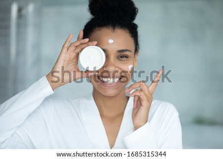 African woman in bathrobe applied cream on face holds jar feels happy looks at camera, anti-wrinkle treatment, remedy for complexion improvement, deep repair, facial skin protection, skincare concept