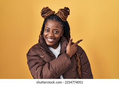 African woman with braided hair standing over yellow background cheerful with a smile on face pointing with hand and finger up to the side with happy and natural expression  - Shutterstock ID 2254774051
