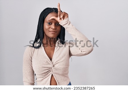 African woman with braids standing over white background making fun of people with fingers on forehead doing loser gesture mocking and insulting. 