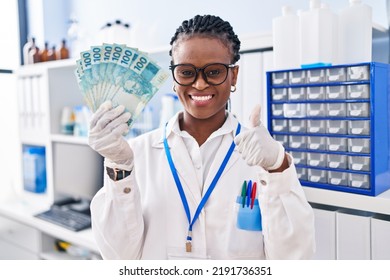 African Woman With Braids Working At Scientist Laboratory Holding Money Smiling Happy And Positive, Thumb Up Doing Excellent And Approval Sign 