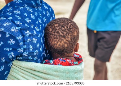 African Woman Carry Her Child Wrapped In A Towel In Her Back In A Traditional Way, People Walking By