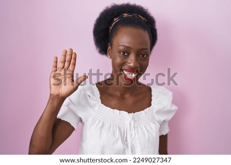 African woman with curly hair standing over pink background waiving saying hello happy and smiling, friendly welcome gesture 