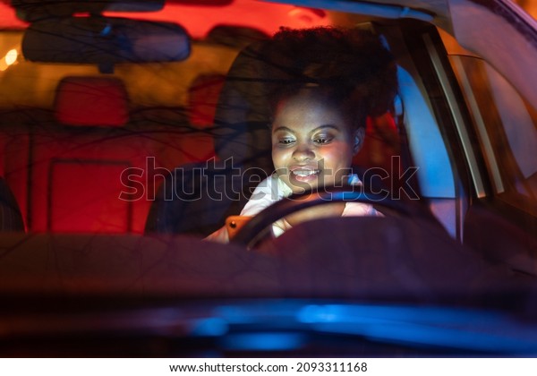 African woman distracted on smart phone while\
driving car at night. Smiling black female read text on mobile\
phone sitting on driver seat. Irresponsible girl messaging riding\
vehicle. Safety on\
road.