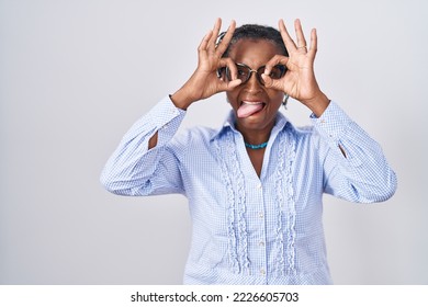 African Woman With Dreadlocks Standing Over White Background Wearing Glasses Doing Ok Gesture Like Binoculars Sticking Tongue Out, Eyes Looking Through Fingers. Crazy Expression. 