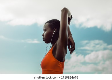 African Woman With Earphones Doing Hand Stretching Exercise Outdoors. Female In Sportswear Doing Warmup Workout Outdoors Against Sky.