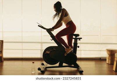 African woman engaging in indoor cycling on a stationary bike, using the smart equipment to follow a cardio routine. Focused and determined, she pedals in pursuit of her health and wellness goals. - Powered by Shutterstock