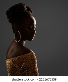African Woman Face Profile Silhouette. Dark Skin Beauty Model with Gold Ring Earrings and Afro Braids Hairstyle in Glitter Golden Dress over Gray Background in Backlight