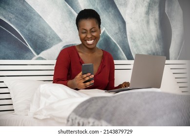 African Woman Freelancer Working From Her Bedroom At Home. Smiling Looking At Her Phone With Laptop, Laying In Bed