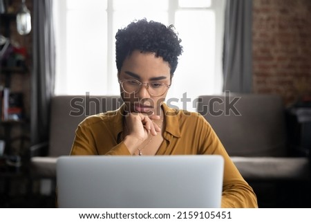 African woman in glasses sit at desk staring at laptop screen, learn new software, makes assignment, working looks concentrated, search solution or ideas. Business challenge, thinks over task concept Foto d'archivio © 