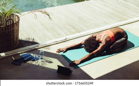 African woman going yoga outdoors by a swimming pool. Fitness woman practicing child pose yoga on exercise mat at the poolside with a digital tablet in front.