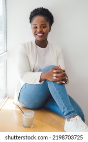 African Woman In Jacket Sitting On Window Sill, Relaxing In Coffee Shop With A Glass Of Iced Coffee. Relaxation Or Weekend Away From Home.