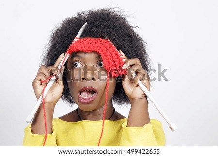 African woman knitting something with red colour, amazed and funny, putting the needles and the yarn on her face. Over a grey background.