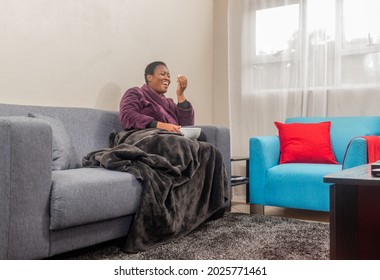 African Woman Laughing While Watching TV, Covered With A Blanket And Eating Pop Corn During Winter Season