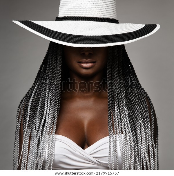 African Woman with Long Braids Hair. Black and\
White Concept. Beauty Model in Big Hat hidden Face. Afro Hairstyle\
and Lips Makeup. Sexy Mysterious Women Portrait over Gray Studio\
Background