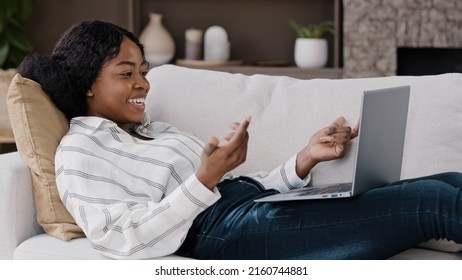 African Woman Lying On Sofa At Home Look At Laptop Screen Greeting Hello Make Video Call Talking Having Remote Conversation With Friend Family Living Abroad. American Female Communicates By Conference