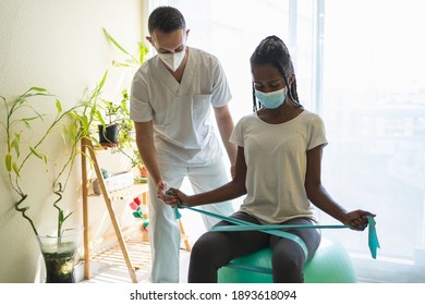 African Woman With Mask Doing Rehabilitation Exercises On Her Legs With An Elastic Band And A Medicine Ball With The Help Of A Physiotherapist In Uniform