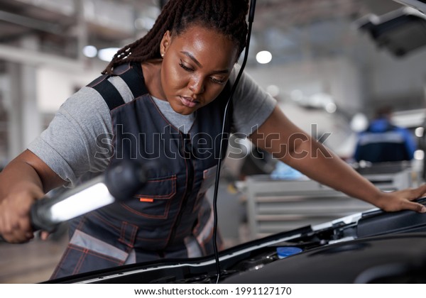 african woman mechanic looking to car engine and\
holding lamp, Car master in service shop, female mechanic repairer\
service technician checks and repairs the engine condition under\
hood of vehicle