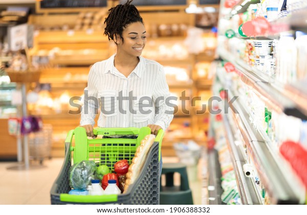 African woman on\
grocery shopping in supermarket pushing cart walking along shelves\
buying food and goods in store. Female customer choosing groceries\
in local shop. Empty\
space