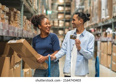 African Woman Picks Up A Box Of Furniture She Has Bought And An Asian Man, Her Boyfriend Holding A Credit Card, Is Comfortable To Use In A Wholesale Store Warehouse.