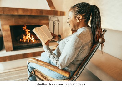 African woman reading book in front of cozy fireplace at home - Autumn and winter lifestyle concept - Focus on ear - Powered by Shutterstock