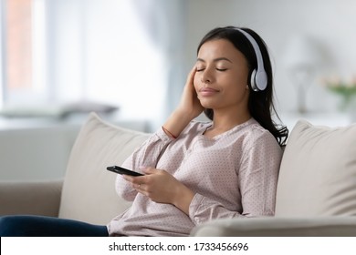 African woman rest leaned on couch in cozy living room spend free time at home enjoy favourite song. Biracial girl wears headphones listen classical music, mood and hobby, modern wireless tech concept - Powered by Shutterstock