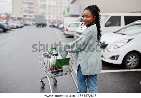 African woman with shopping cart trolley posed\
outdoor market near car\
parking.