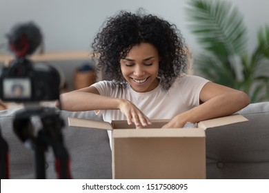 African woman sit on sofa captures on SLR or DSLR digital camera moment of unpacking ordered goods on internet to share product feedback with subscribers, vlogger filming material for channel concept