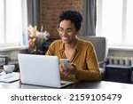 African woman sit at workplace desk holds cellphone staring at laptop, synchronize data between computer and gadget in office, use corporate devices and business application, plan work, use organizer