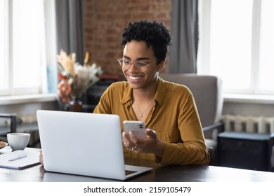 African woman sit at workplace desk holds cellphone staring at laptop, synchronize data between computer and gadget in office, use corporate devices and business application, plan work, use organizer