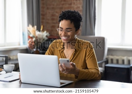 African woman sit at workplace desk holds cellphone staring at laptop, synchronize data between computer and gadget in office, use corporate devices and business application, plan work, use organizer