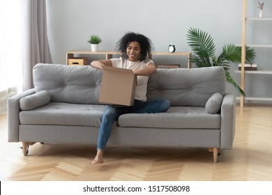 African woman sitting on couch in living room hold on lap big carton box unpacking received parcel feels happy, ordered online goods quick shipped at home, transport and delivery services ad concept
